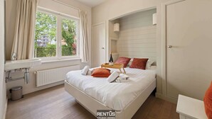 Une chambre à coucher avec un lit, une fenêtre avec des rideaux, une table de chevet avec une bouteille et des verres, et un radiateur.