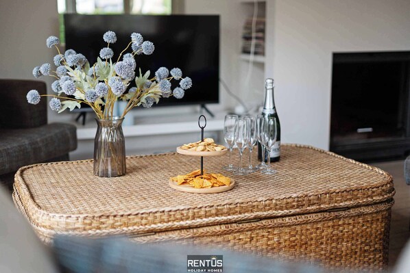 A living room with a TV, a fireplace, a vase with flowers, and a wicker table with snacks and glasses.