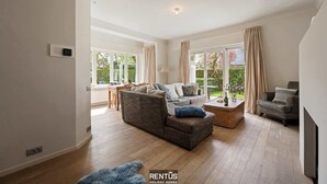 Un salon spacieux avec un canapé, un pouf et une table basse. De grandes fenêtres sont agrémentées de rideaux, et on y trouve une lampe ainsi qu’une cheminée.