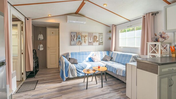 A cozy room with a striped sofa, a small round table, a kitchenette, and a window with curtains. - Mh 229 - La Tremblade: Comfortable Mobile Home for Families, Near Beach & City Center (La Tremblade)