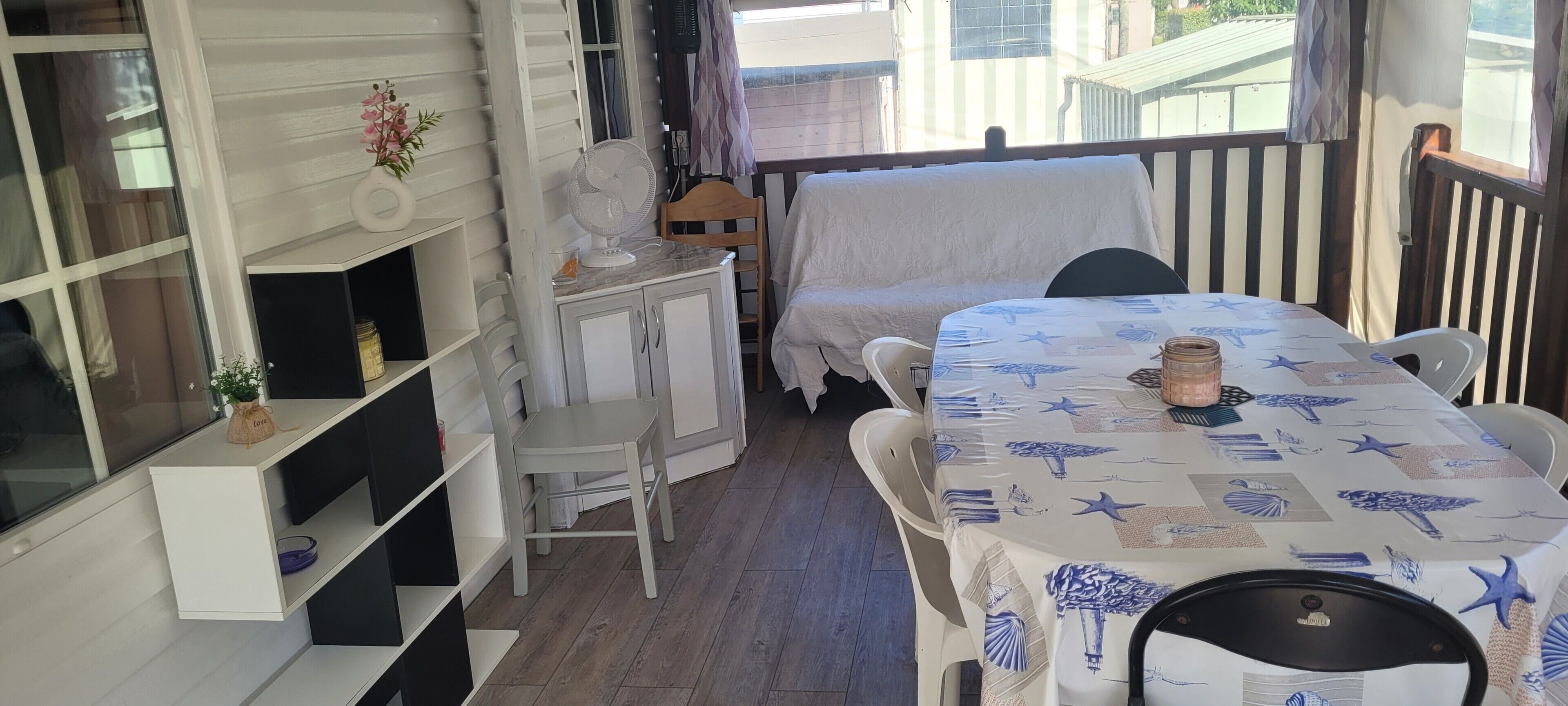 Una habitación con una cama, una mesa con mantel de estrella de mar, un ventilador y una repisa con una planta pequeña.