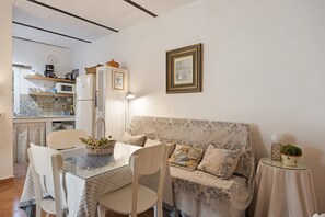 A cozy living area with a sofa, a dining table with chairs, a kitchen in the background, and a small side table with a plant.