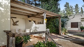Un área de estar al aire libre techada con sillas y una mesa, rodeada de plantas en macetas y una valla de madera.
