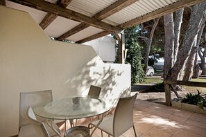 A covered patio with a round glass table and four chairs, surrounded by trees and a white wall.