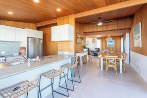 A modern kitchen with wooden cabinetry, a stainless steel refrigerator, bar stools, and a dining area with a table and chairs.