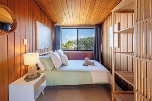 Un dormitorio con techo de madera, un ventanal grande, una cama con almohadas, una mesita de noche con lámpara y un armario.