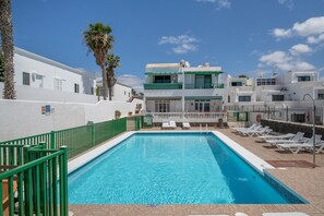Una piscina con sillas de descanso, un edificio blanco con balcones y palmeras.