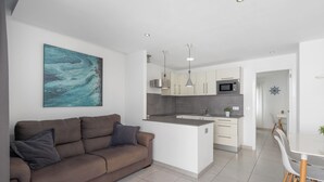 A modern living room with a sofa, a dining table with chairs, a kitchen area with cabinets and appliances, and a large abstract painting on the wall. - Vacation 'Princesa Teguise' with Shared Pool, Shared Terrace and Wi-Fi (Tías)