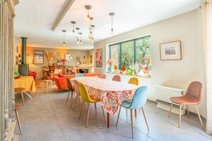 Un coin repas avec une table recouverte d’une nappe à motifs floraux, entourée de chaises colorées, une grande fenêtre donnant sur des arbres et un radiateur au mur.