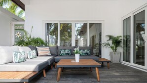 Un área de estar exterior moderna con un sofá gris, una mesa de centro de madera y cojines con motivos tropicales.