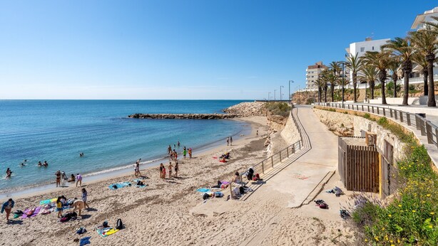 Una playa con gente tomando el sol y nadando, una orilla arenosa y edificios al fondo.