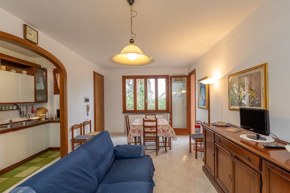 A living room with a sofa, a dining table with chairs, a kitchen area, and a television on a wooden cabinet.