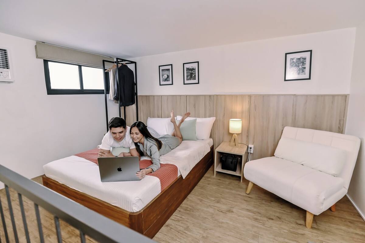 A modern hotel room with a wooden bed, a white sofa, and a nightstand with a lamp.