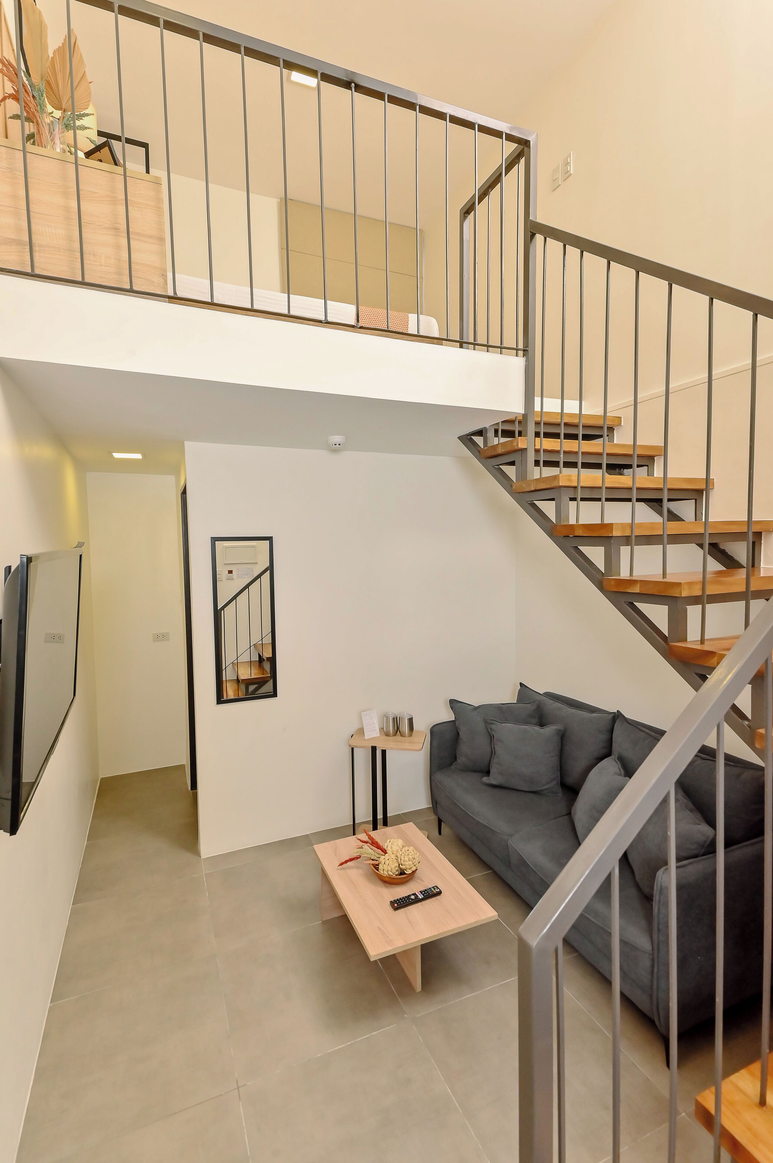 A modern apartment with a staircase, a grey sofa, a wooden coffee table, and a small table with a vase and remote control.
