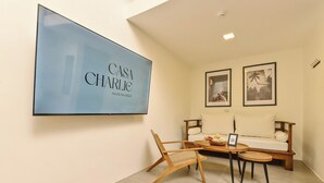 A modern living room with a flat-screen TV displaying 'Casa Charlie' branding, a wooden sofa, a wicker chair, and a small round table with a bowl and a framed picture.