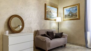 Een kamer met een beige bank, een witte kapstok, een ronde spiegel en twee ingelijste schilderijen aan de muur.