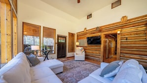 Ein gemütliches Blockhaus-Interieur mit einem weißen Ecksofa, einem kleinen runden Tisch mit Lampe und einem an der Wand befestigten Fernseher.