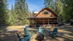 Eine Hütte mit überdachter Veranda, umgeben von Bäumen. Vier Adirondack-Stühle und eine Feuerstelle befinden sich im Garten.