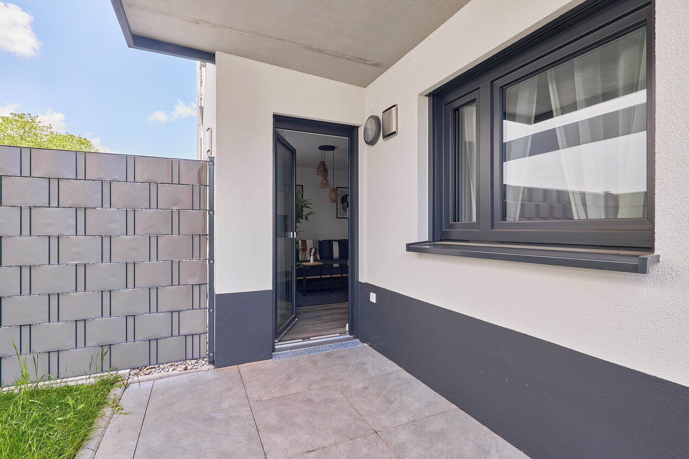 A modern exterior with a grey brick wall, a sliding glass door leading to an interior with a sofa and pendant lights, and a window with a white curtain.