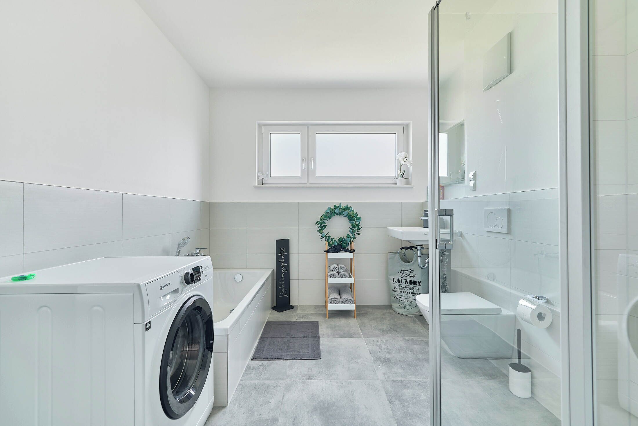 Une salle de bain moderne équipée d’un lave-linge, d’une baignoire, de toilettes et d’une douche à parois de verre.