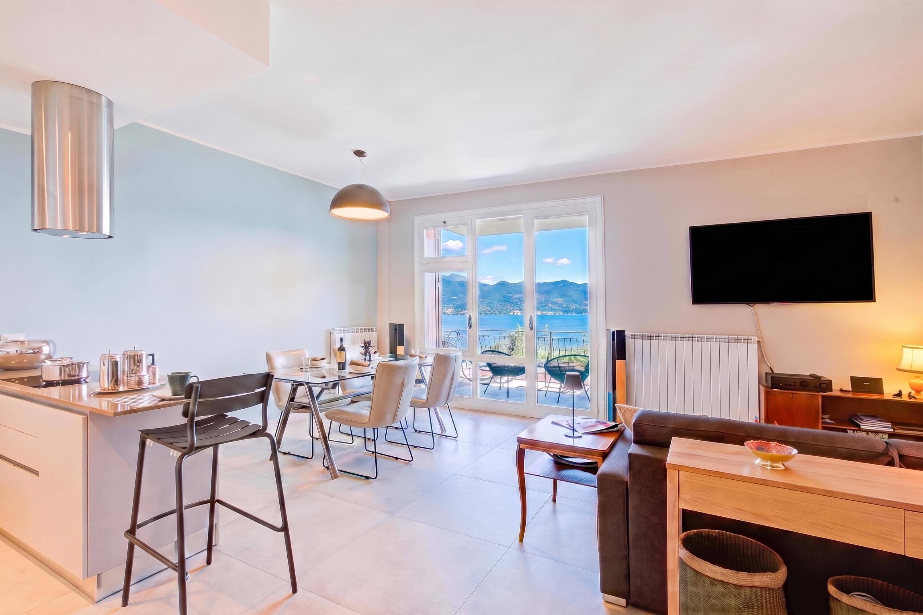 A modern kitchen with a dining area, a flat-screen TV mounted on the wall, and a balcony with a view of the sea.