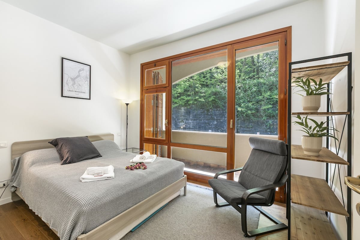 A bedroom with a bed, a chair, a window, and a shelf with plants.