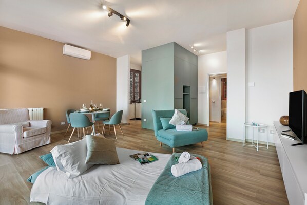 A modern hotel room with a bed, a sofa, a dining area, and a TV.