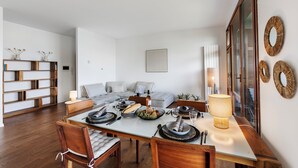 Un coin repas avec une table en bois dressée pour quatre personnes, entourée de chaises. On aperçoit un canapé au fond et une bibliothèque sur la gauche.