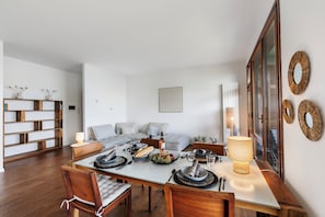 Un coin repas avec une table en bois dressée pour quatre personnes, entourée de chaises. On aperçoit un canapé au fond et une bibliothèque sur la gauche.