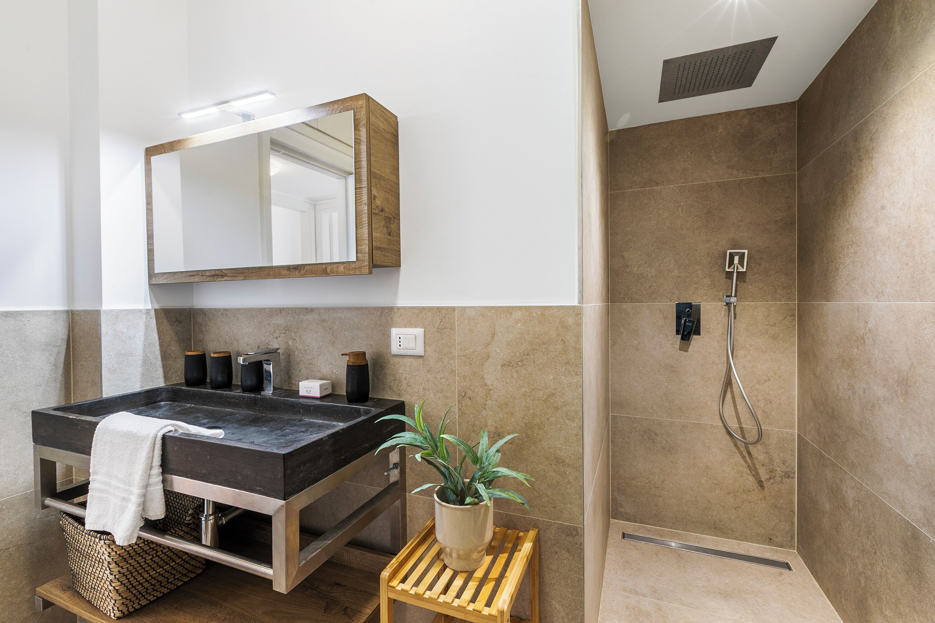 A modern bathroom with a large dark sink, a mirror, a shower, and a small wooden stool with a plant.