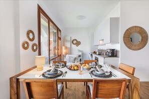 Un coin repas avec une table en bois dressée pour quatre personnes, entourée de chaises. On aperçoit une cuisine au fond.