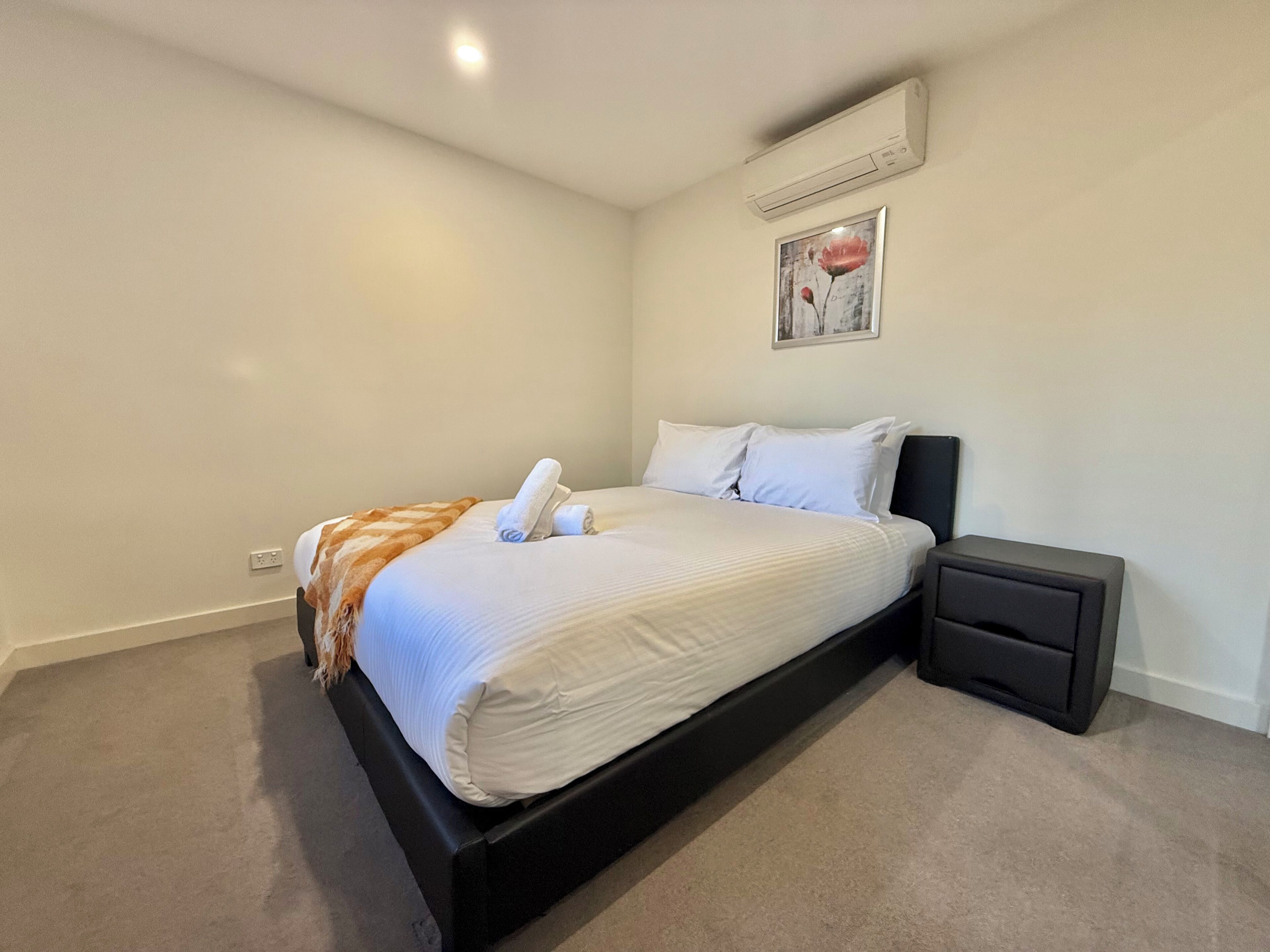 A bedroom with a bed, a bedside table, a wall-mounted air conditioner, and a framed picture on the wall.
