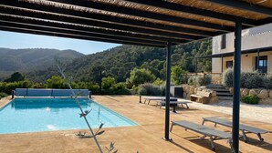 A pool with a pergola overhead, outdoor seating, and a view of a mountainous landscape.