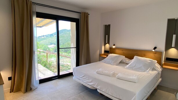A hotel room with a bed, a large sliding glass door to a balcony, and a view of a green landscape.
