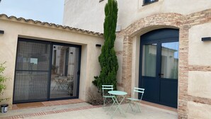 A building with a large sliding glass door, a tall green tree, and a small patio with two chairs and a table.