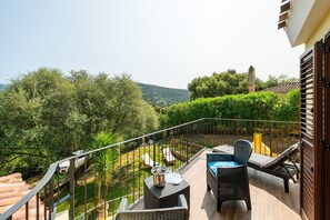 Un balcon agrémenté de meubles en osier, d’une table et offrant une vue sur un jardin luxuriant et des collines lointaines.