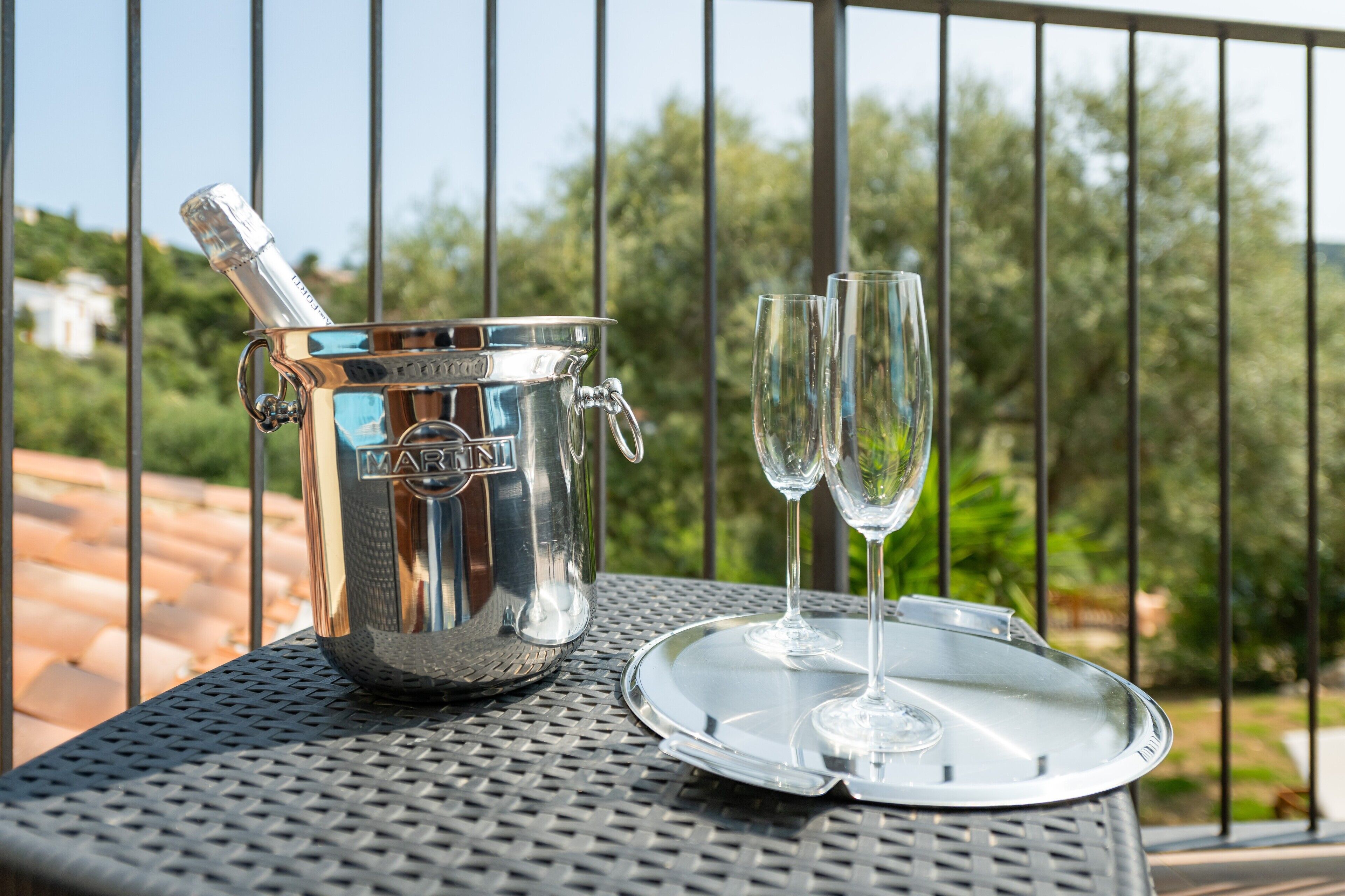 A metallic table with a champagne bucket and two glasses on a tray, set outdoors with a metal railing and greenery in the background.