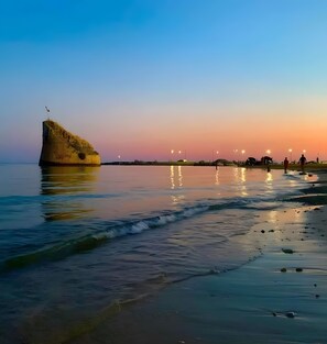 A beach with a large rock formation, calm water, and distant lights. - Holiday Home 'Nicola' with Private Terrace and Air Conditioning (Salve)