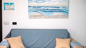 Une pièce avec un canapé bleu, des coussins beige, un tableau représentant une scène de plage accroché au mur, une carte bleue et un thermostat.