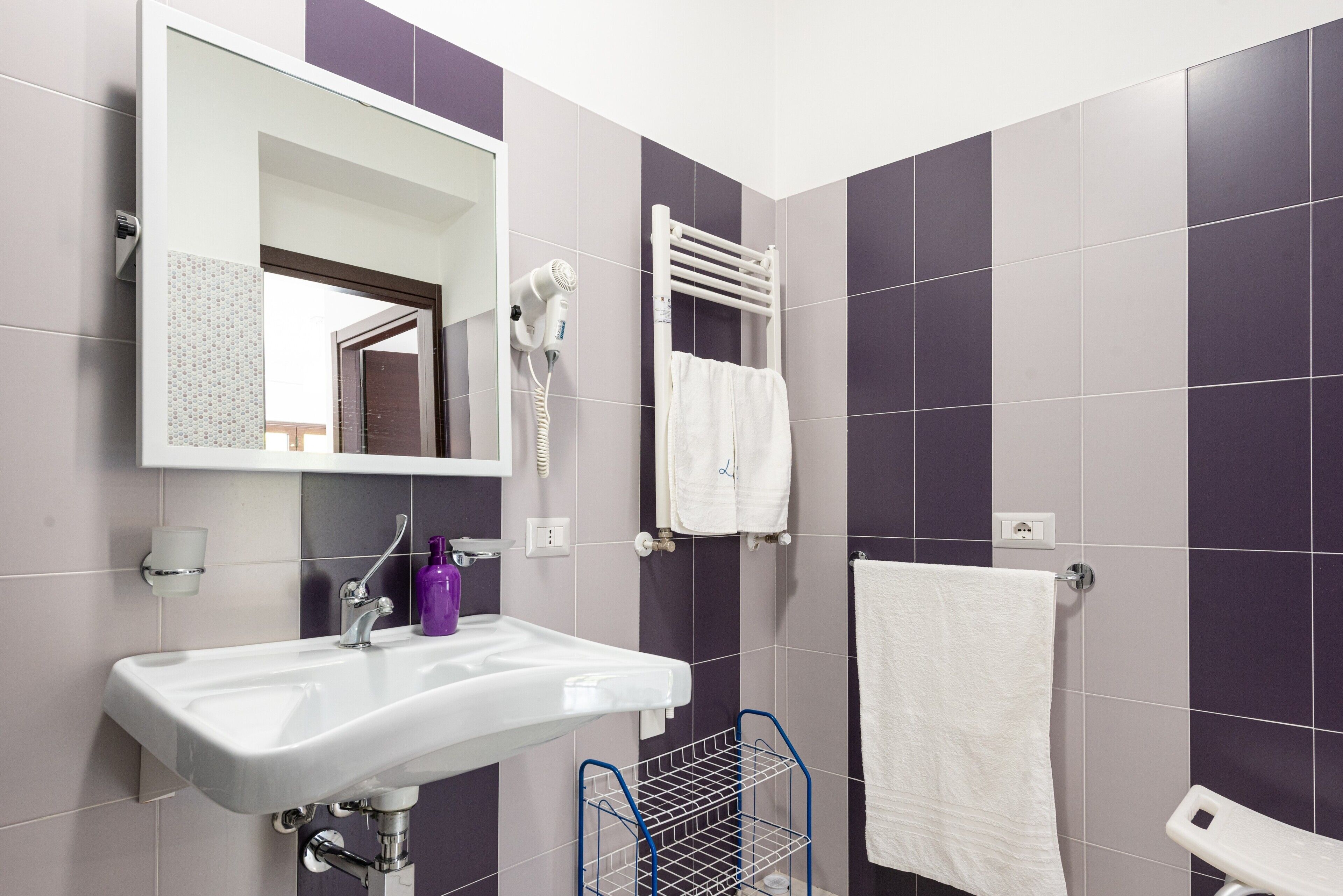 Une salle de bain avec un lavabo blanc, un miroir, un sèche-cheveux, un porte-serviettes et une petite étagère contenant des articles de toilette.