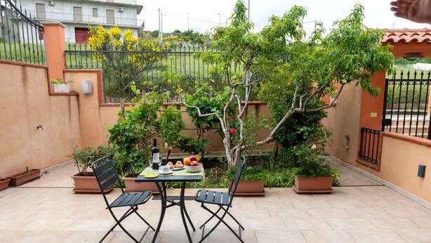 Eine Terrasse im Freien mit Tisch und Stühlen, umgeben von Topfpflanzen und einem Zaun.