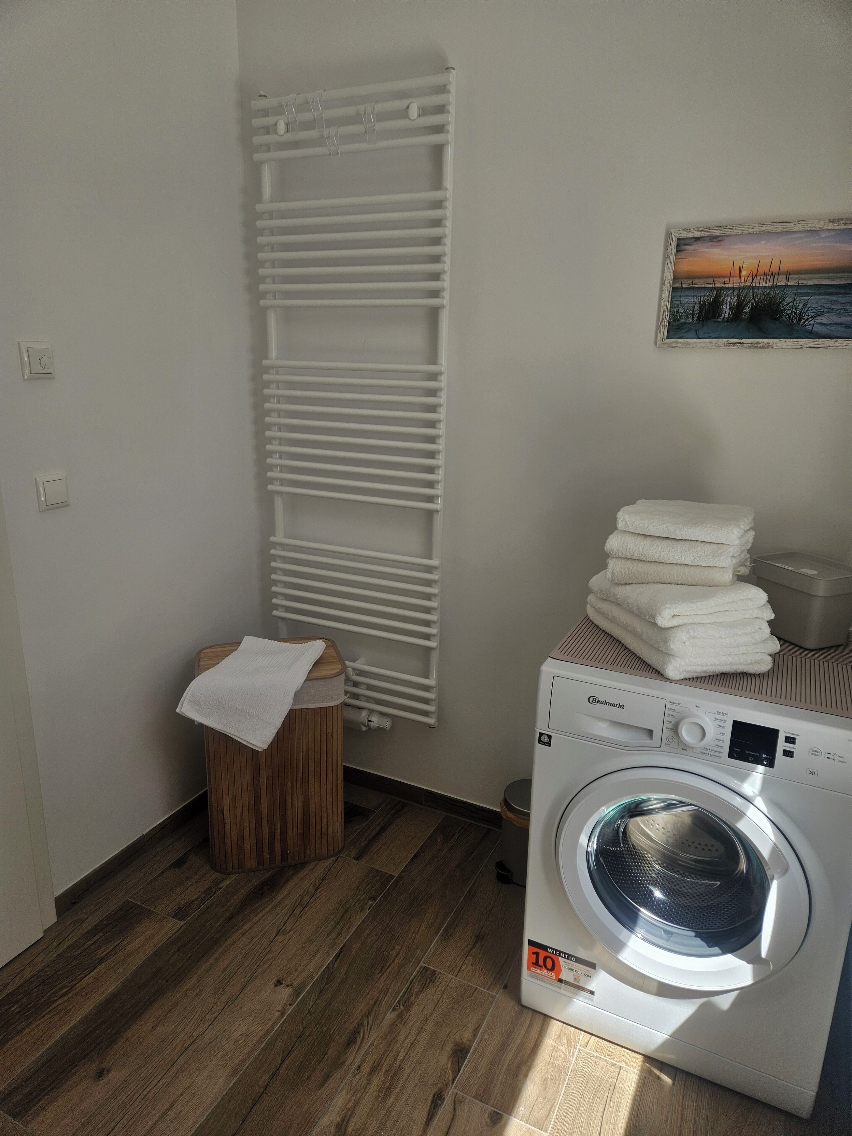 A laundry room with a white radiator, a stack of white towels, a wooden stool, and a front-loading washing machine.