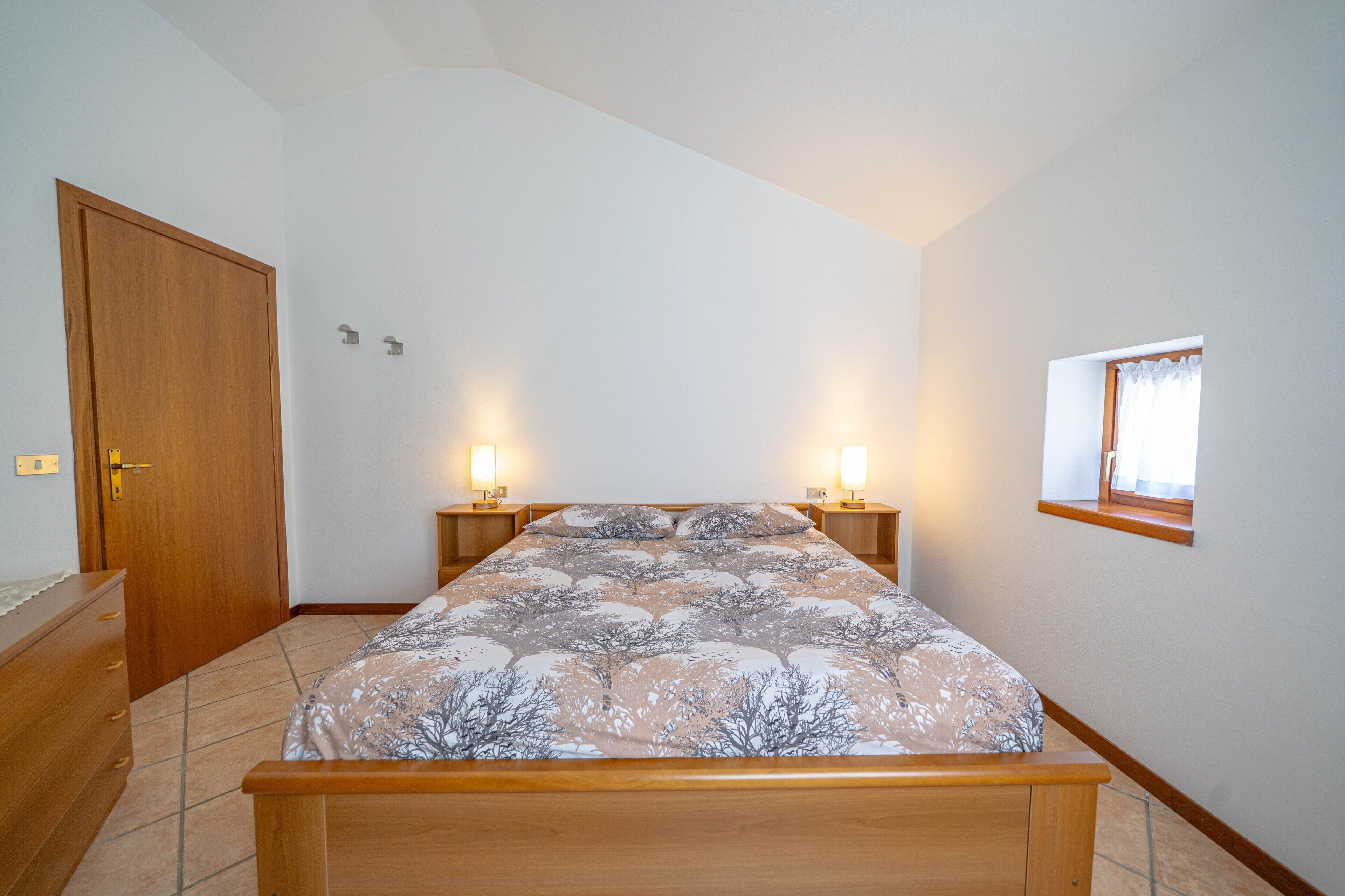 Un dormitorio con cama, cómoda de madera, lámparas de noche y ventana.