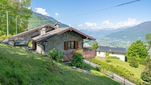 A stone house with a wooden balcony, surrounded by lush greenery and mountains in the background. - Holiday Home 'La Rosa Bianca' with Mountain View, Private Terrace and Wi-Fi (Morbegno)