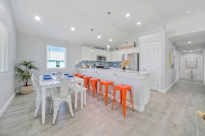A modern kitchen with white cabinetry, a central island with stools, and a dining area with white chairs. - Sleeps 21, 2 Homes | Private Pool, Patio Lounge w/ Fire Pit, Bar, TV, & Games (Port Aransas)