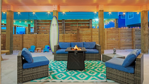 An outdoor seating area with wicker furniture, a fire pit, and a surfboard. - Sleeps 21, 2 Homes | Private Pool, Covered Patio Lounge, Bar, TV, & Games (Port Aransas)