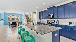 A modern kitchen with blue cabinets, a granite countertop, and a dining area with green stools. - Pool, Hot Tub+Game Rm, Mins to Disney & Universal | Sunshine Ridge by AvantStay (Kissimmee)