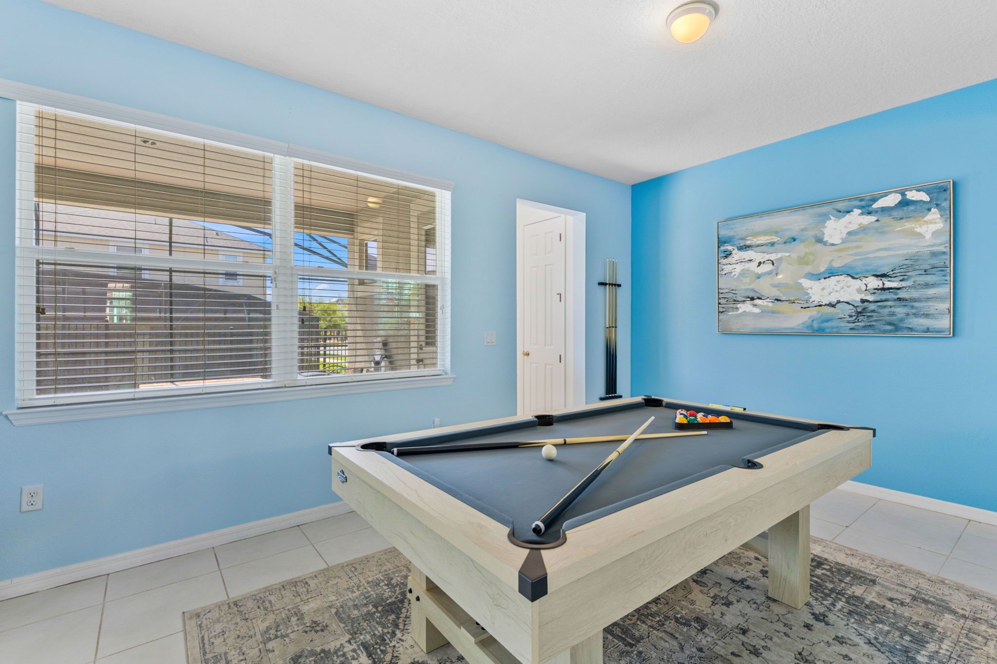 Una mesa de billar en una habitación con paredes azul claro y una ventana con persianas.
