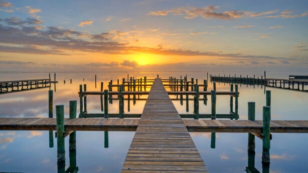 Une jetée en bois s’étendant sur une étendue d’eau calme, avec un coucher de soleil en arrière-plan.