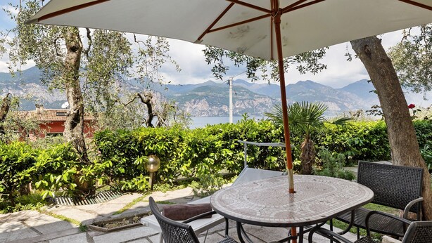 A patio with a round table and chairs, surrounded by greenery and a view of mountains. - Apartment 'Panorama' with Lake View, Wi-Fi and Air Conditioning (Brenzone sul Garda)
